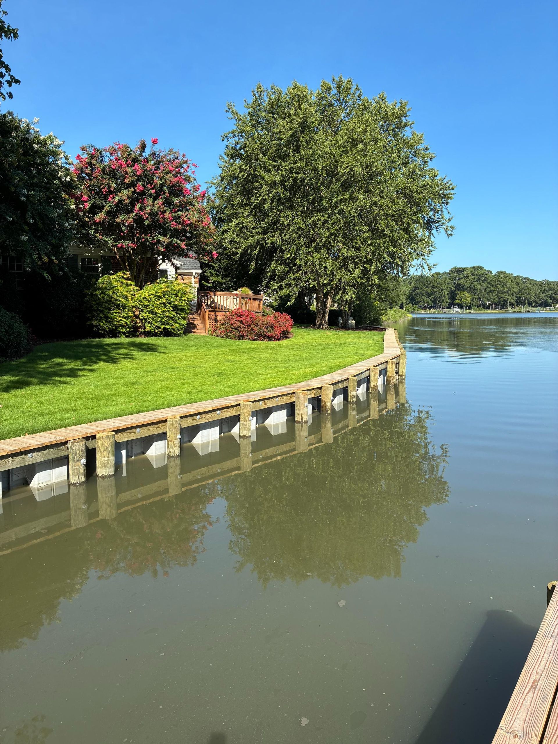 Lush green lawn meets calm water.  Trees and a home border the water's edge, under a clear blue sky.
