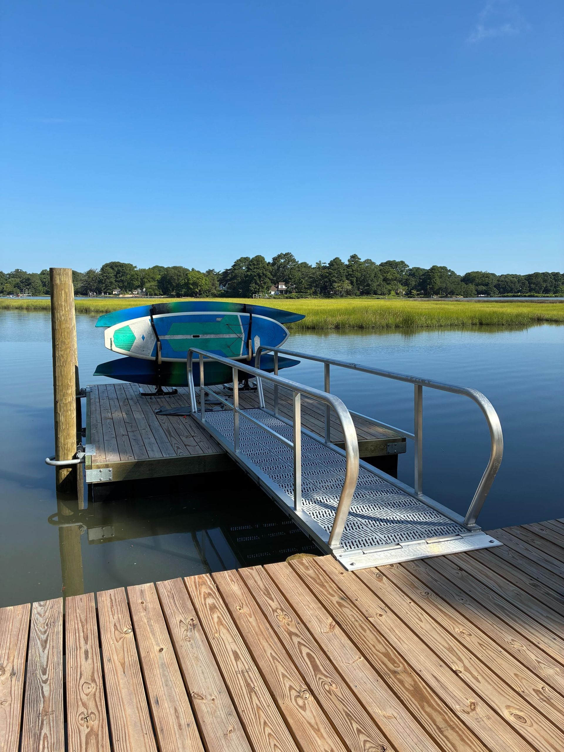 Dock with ramp leading to floating platform holding surfboards, water, and marsh in the background.