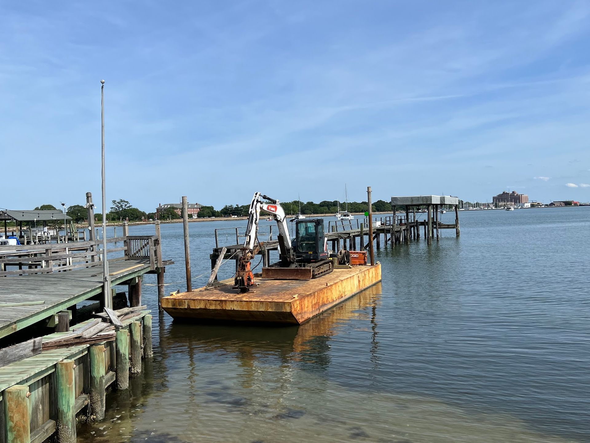 Excavator on a rusty barge in water near a wooden dock under a blue sky.