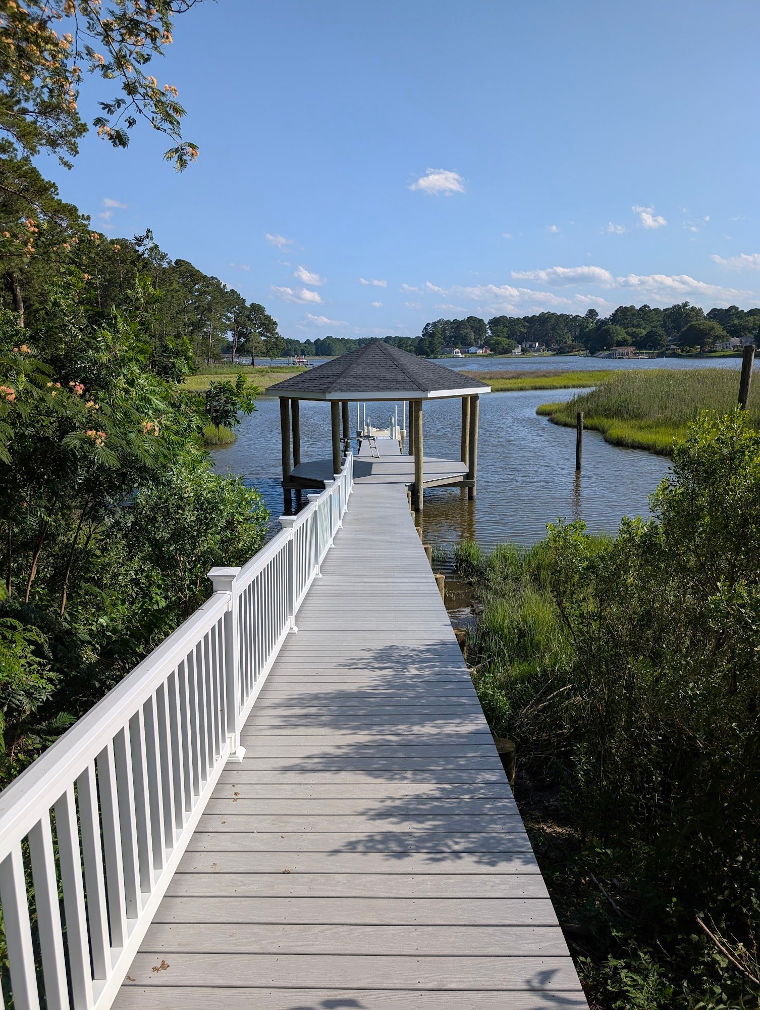 Wooden dock extending to covered gazebo over water, flanked by lush greenery, under blue sky.
