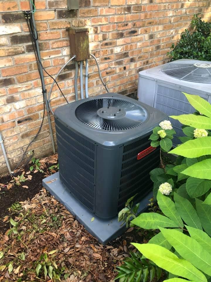 Two outdoor air conditioning units sit on concrete pads against a brick exterior wall next to green foliage.