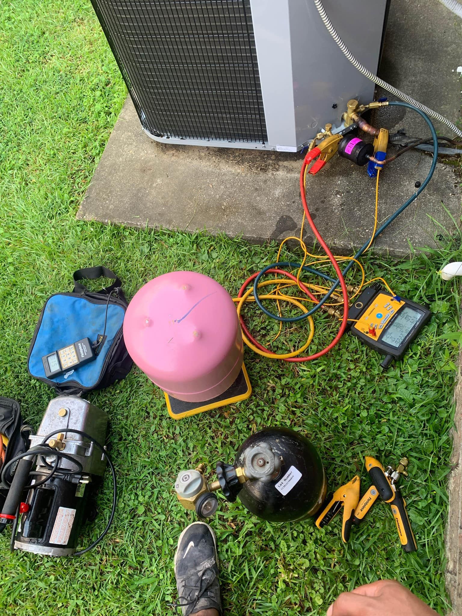 Technician tools for HVAC maintenance, including a refrigerant tank, vacuum pump, and pressure gauges, on a grassy lawn.