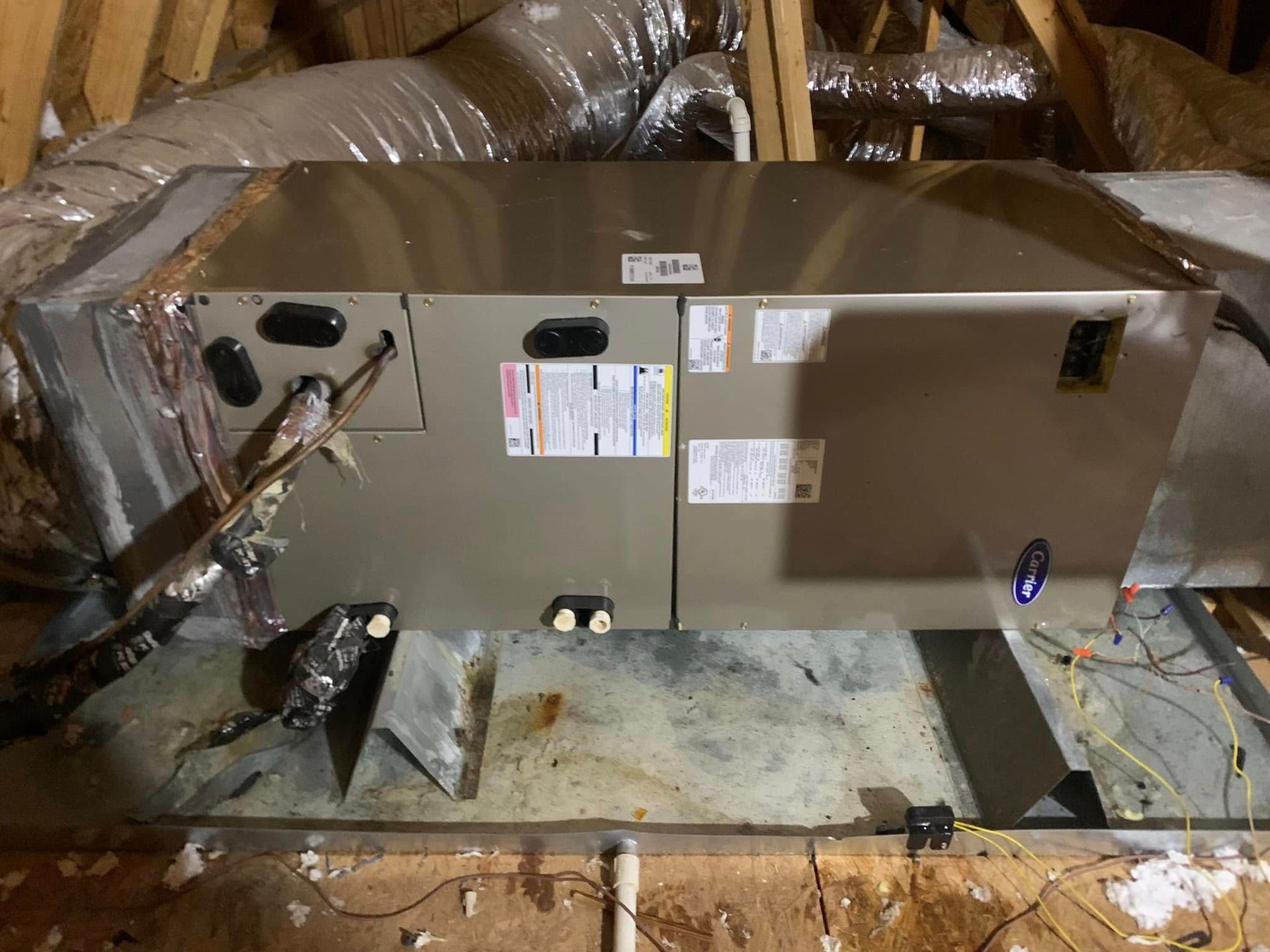 A metal HVAC air handler unit installed in an attic space, with connected insulated ductwork and piping.