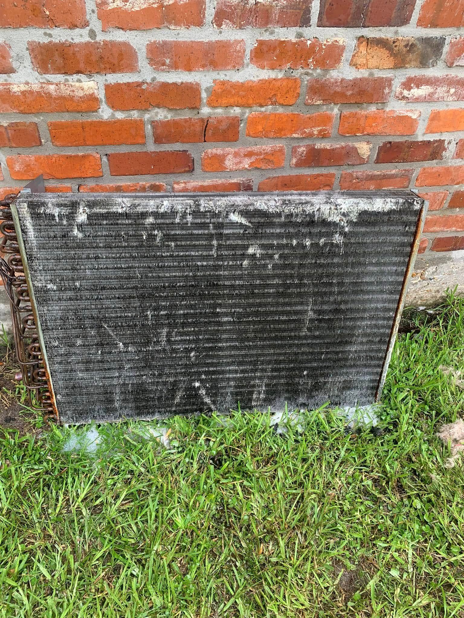 A dirty, rectangular air conditioner radiator coil standing on grass in front of a red brick wall.