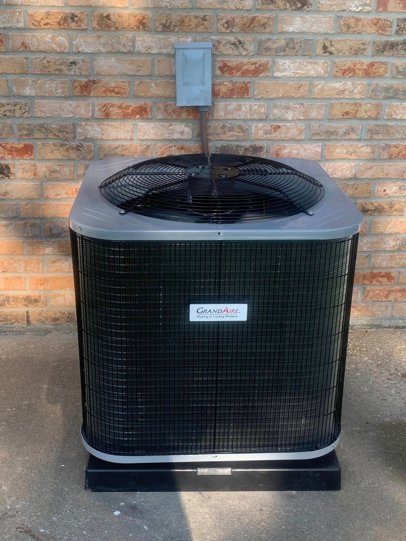 A black central air conditioning unit sits on a concrete pad against a brick wall beneath a mounted electrical disconnect.