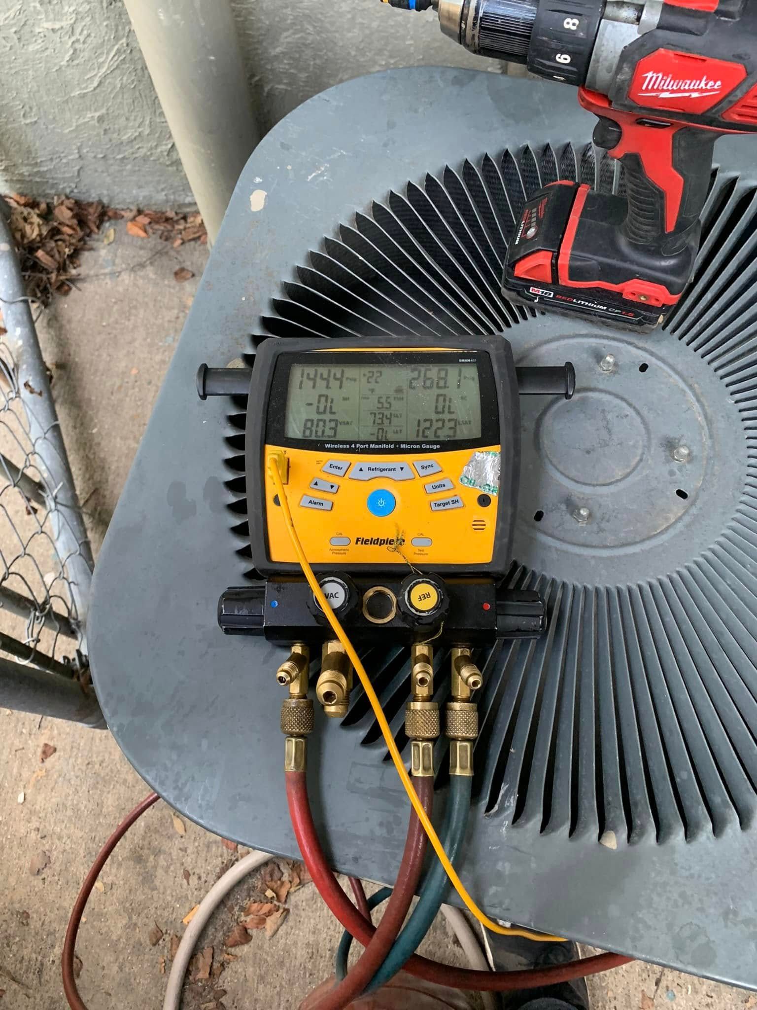 A digital HVAC manifold gauge connected to an outdoor air conditioning unit with a power drill resting on the fan grille.