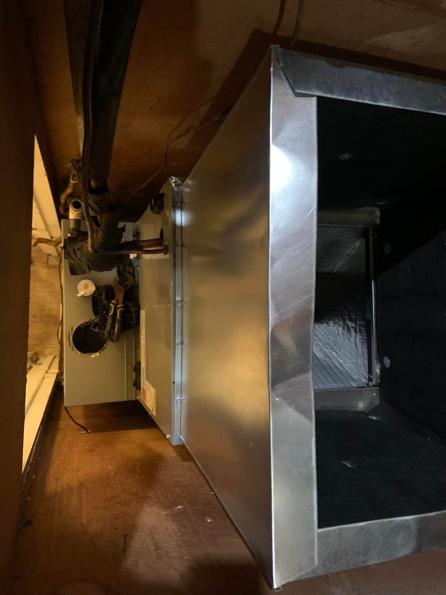 A metal HVAC air handler unit in a utility space, viewed from the side, with pipes and cables visible in the background.