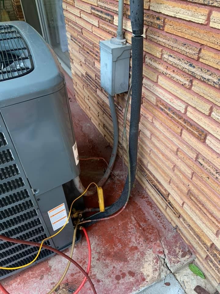 An outdoor air conditioner unit connected to a gray electrical box on a brick wall, with refrigerant hoses attached.