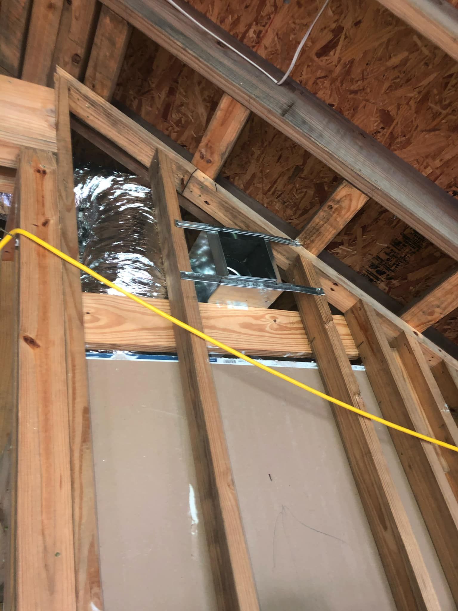 A metal HVAC air duct boot is mounted between exposed wooden wall studs and ceiling rafters in an unfinished room.
