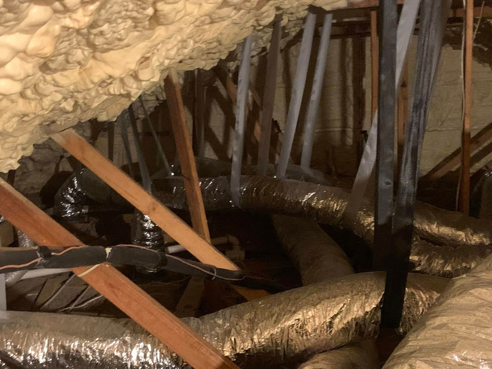Attic view showing spray foam insulation on the roof decking and silver flexible air ducts between wooden rafters.