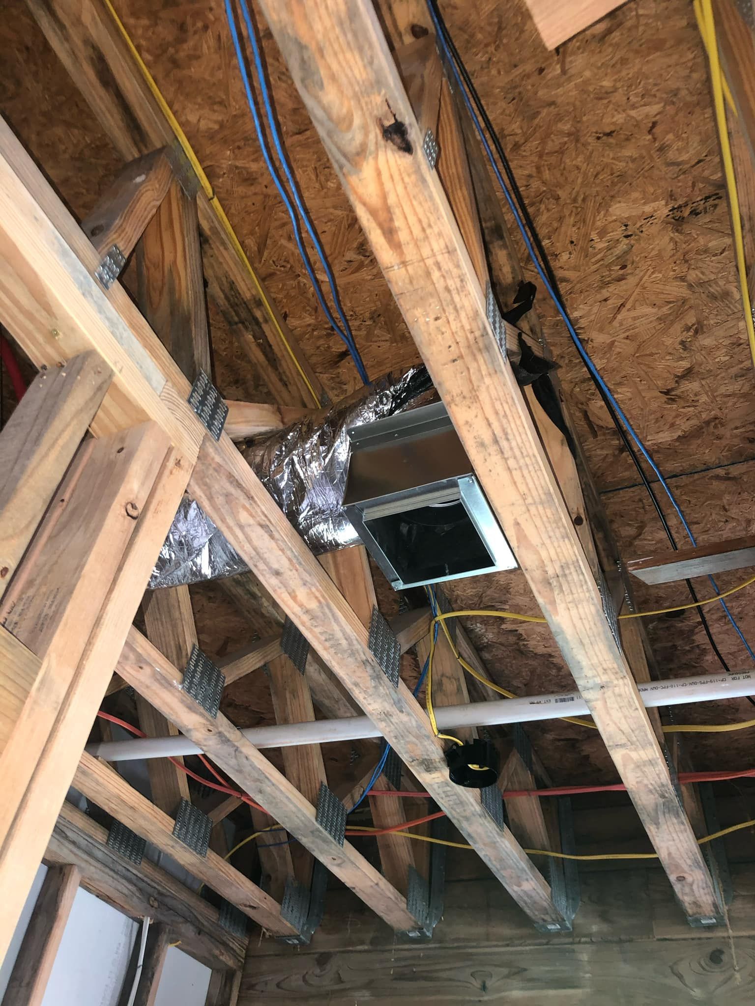 An unfinished attic framing with exposed wooden trusses, HVAC ductwork, and various electrical cables and pipes.