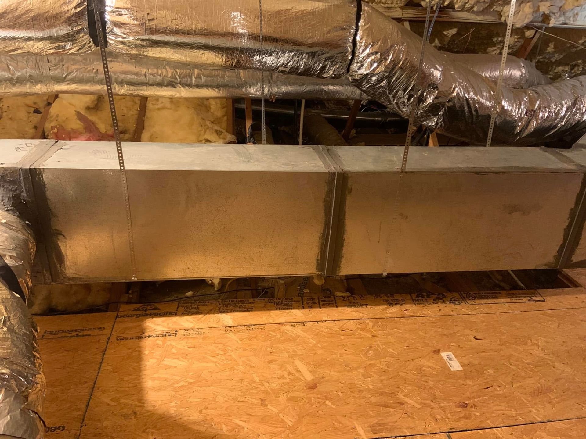 A rectangular metal HVAC duct suspended by chains in an attic, with flexible silver ductwork visible in the background.