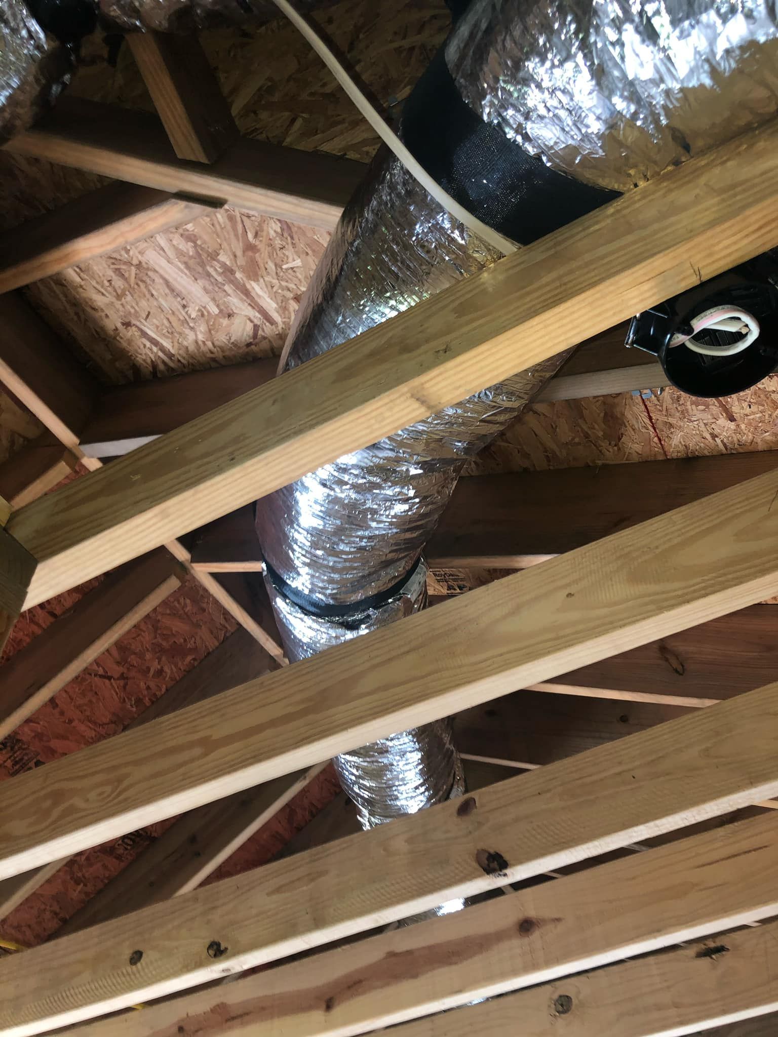 Flexible silver HVAC ducting running through wooden roof trusses in an attic space.