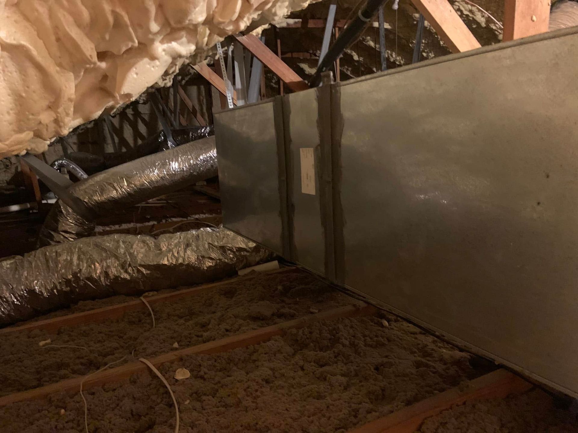 A rectangular metal duct runs through an attic space, with flexible ductwork and spray foam insulation visible nearby.