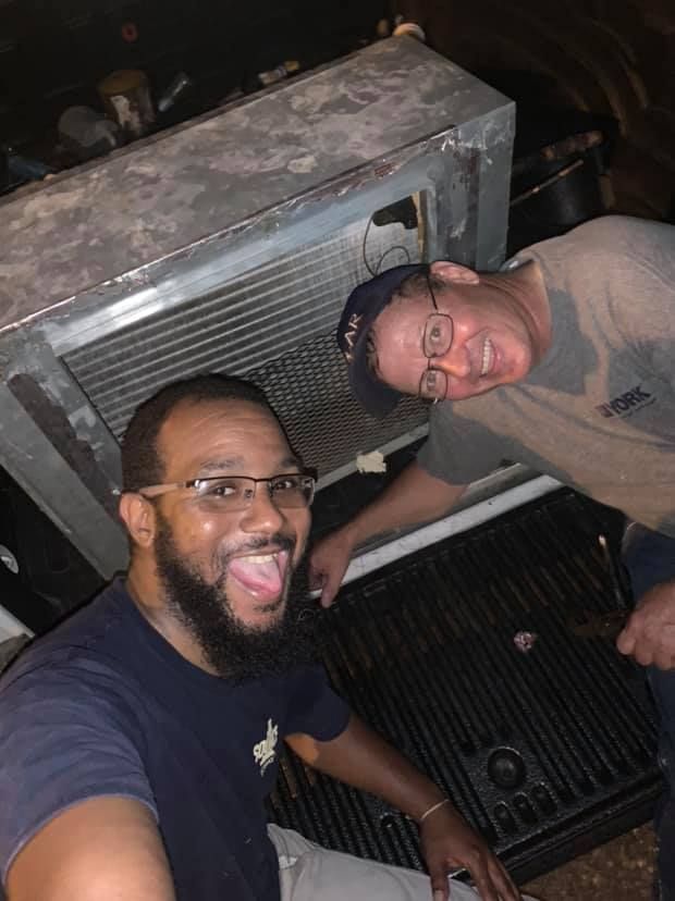 Two people wearing glasses working on a large metal mechanical unit in an outdoor or industrial setting.