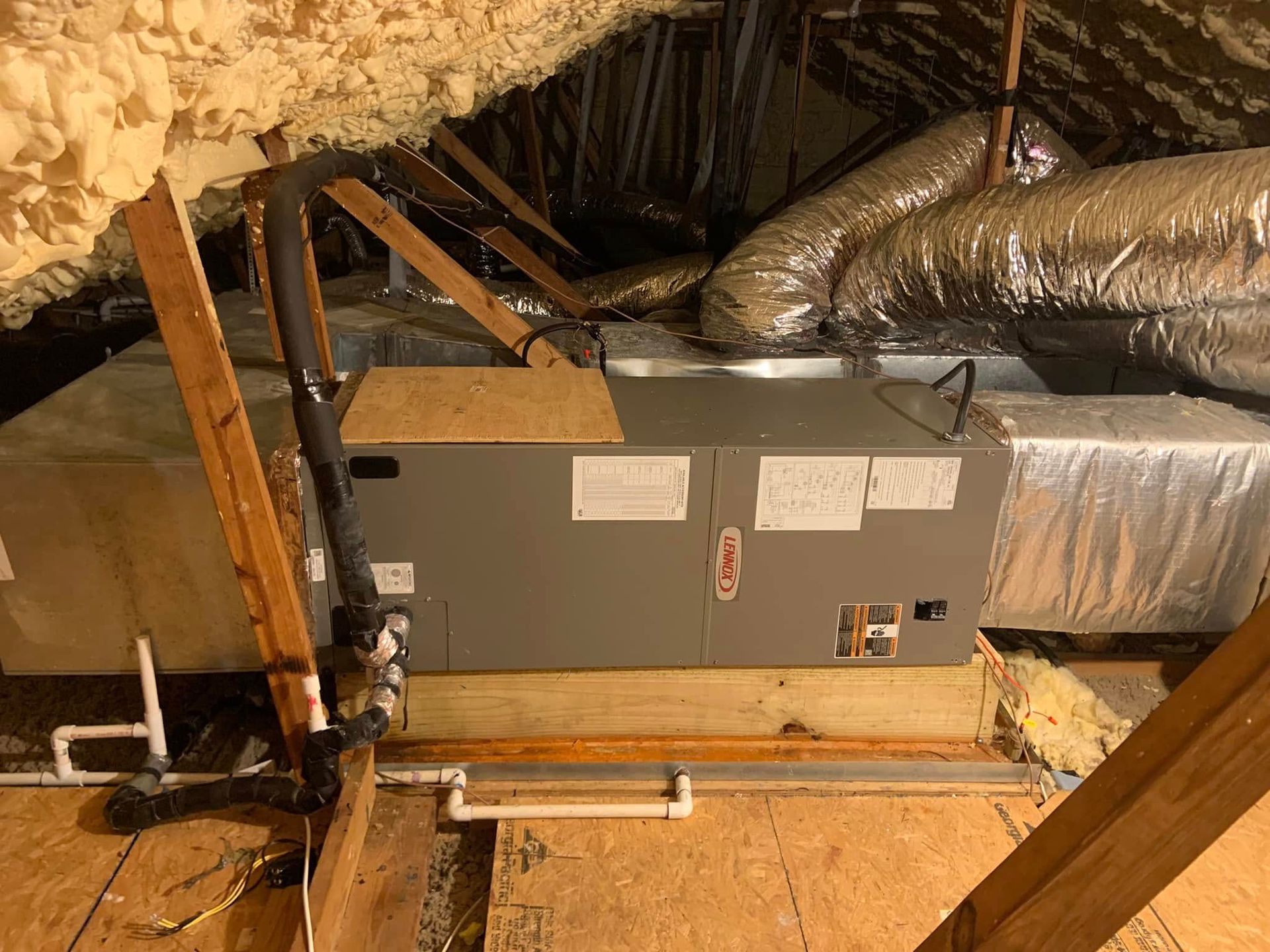 An HVAC air handler unit installed in an attic with spray foam insulation, silver ductwork, and white drainage pipes.