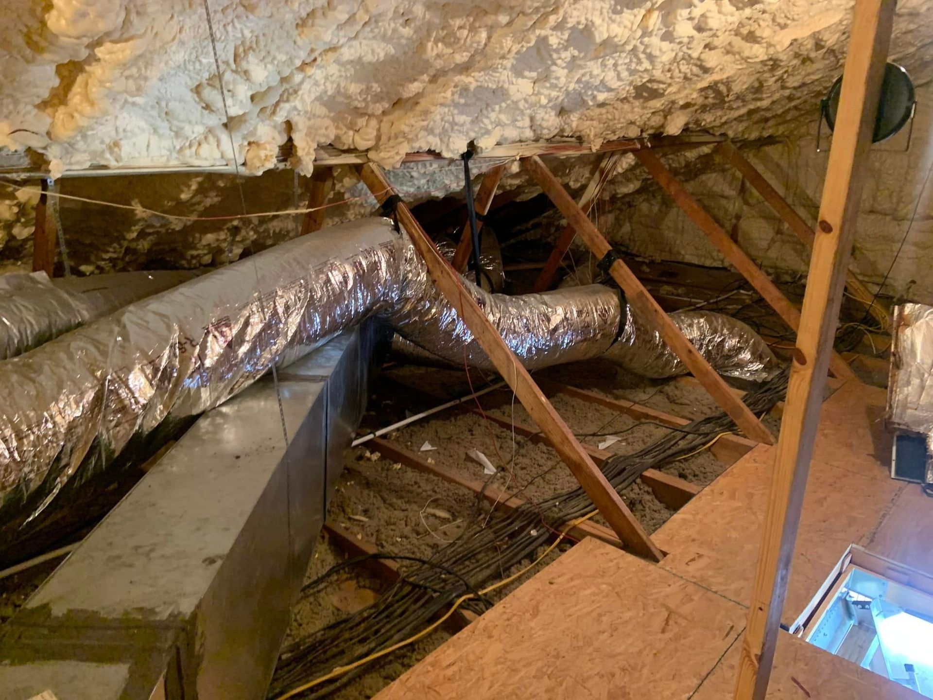 Attic interior with spray foam insulation on the ceiling, metallic HVAC ductwork, and wooden roof trusses.