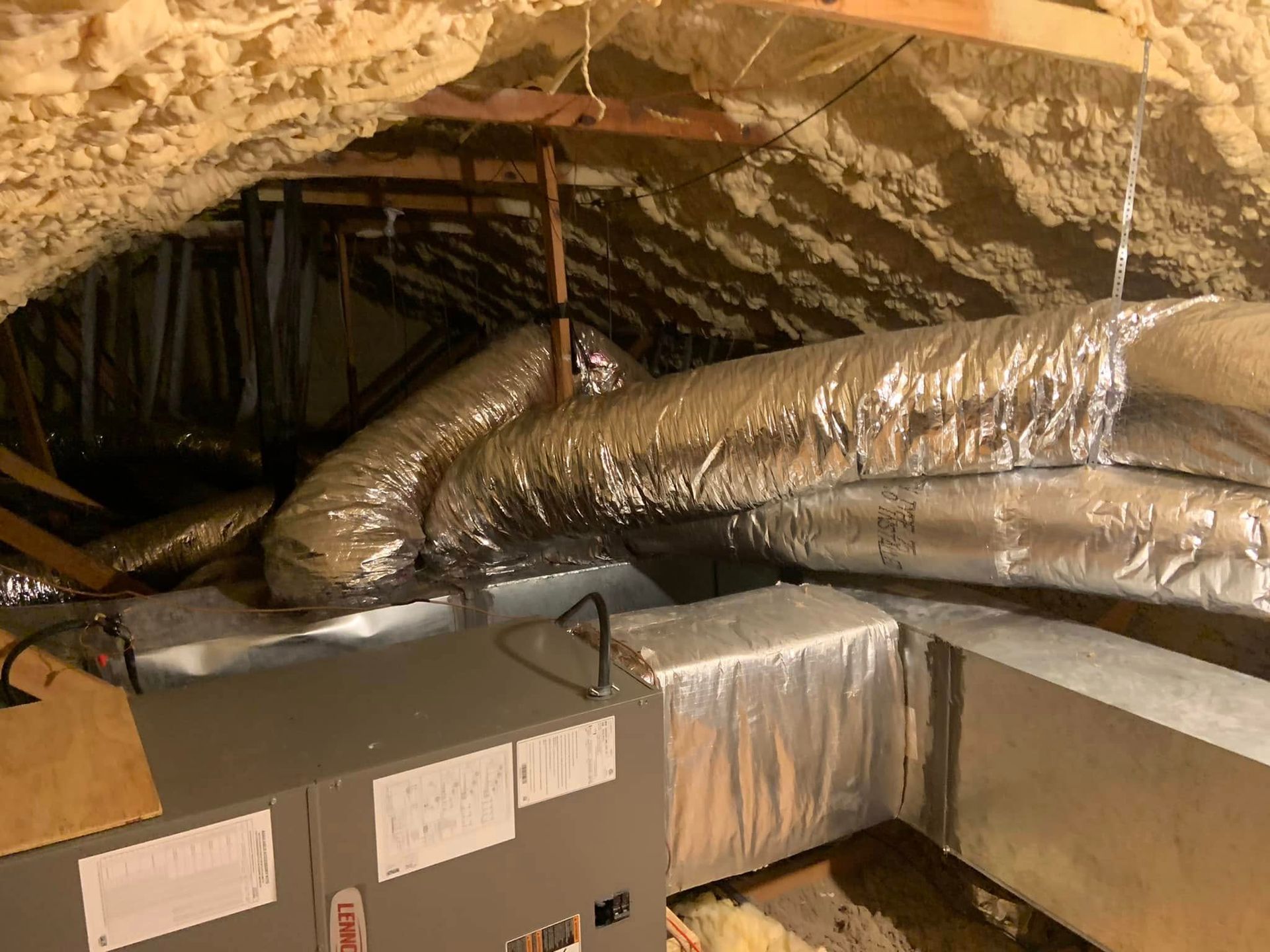 A gray Lennox HVAC air handler unit connected to metallic ductwork in an attic with spray-foam insulated roof rafters.