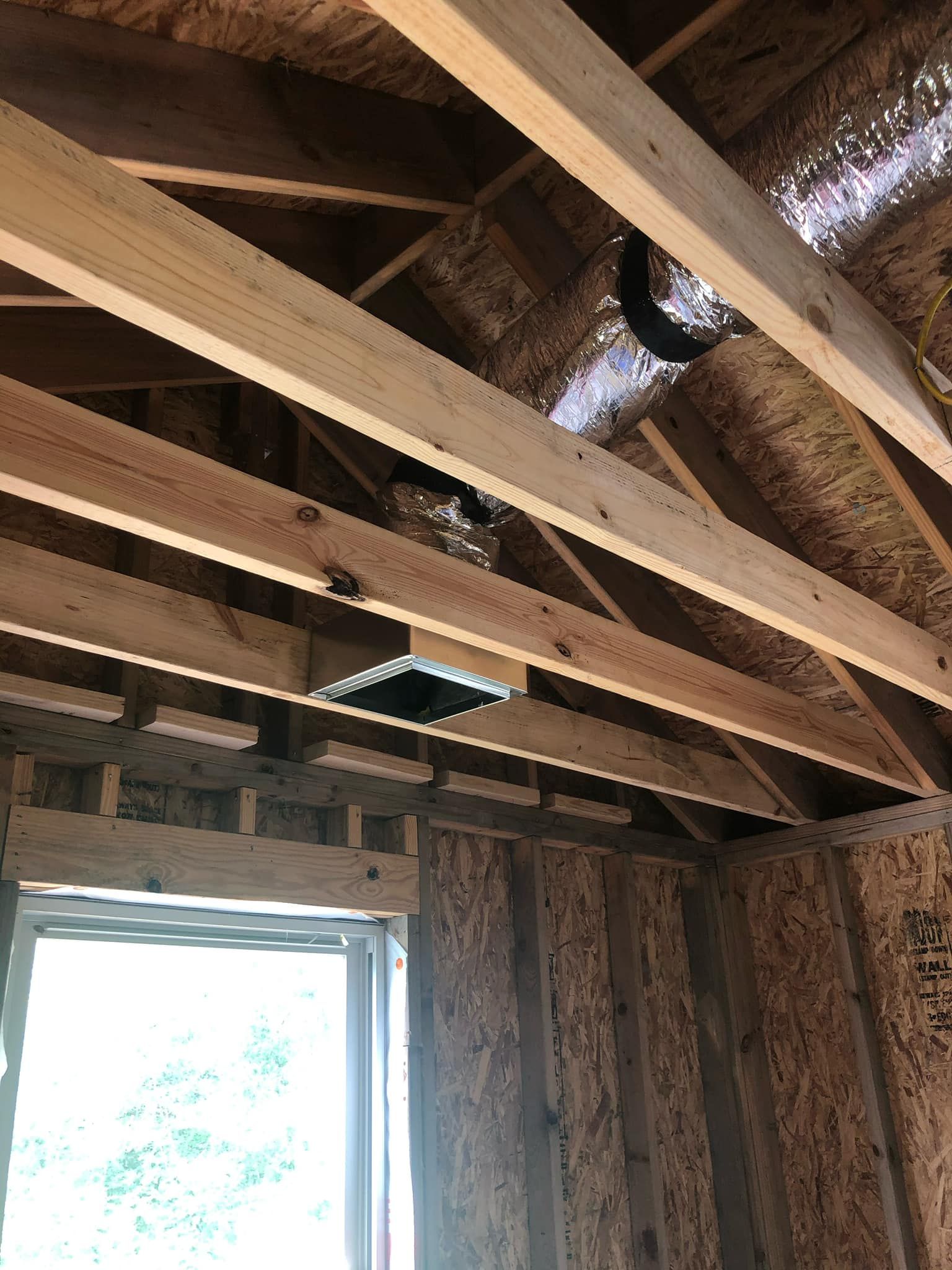 A square ventilation register installed between wooden ceiling joists with metallic ductwork visible above.