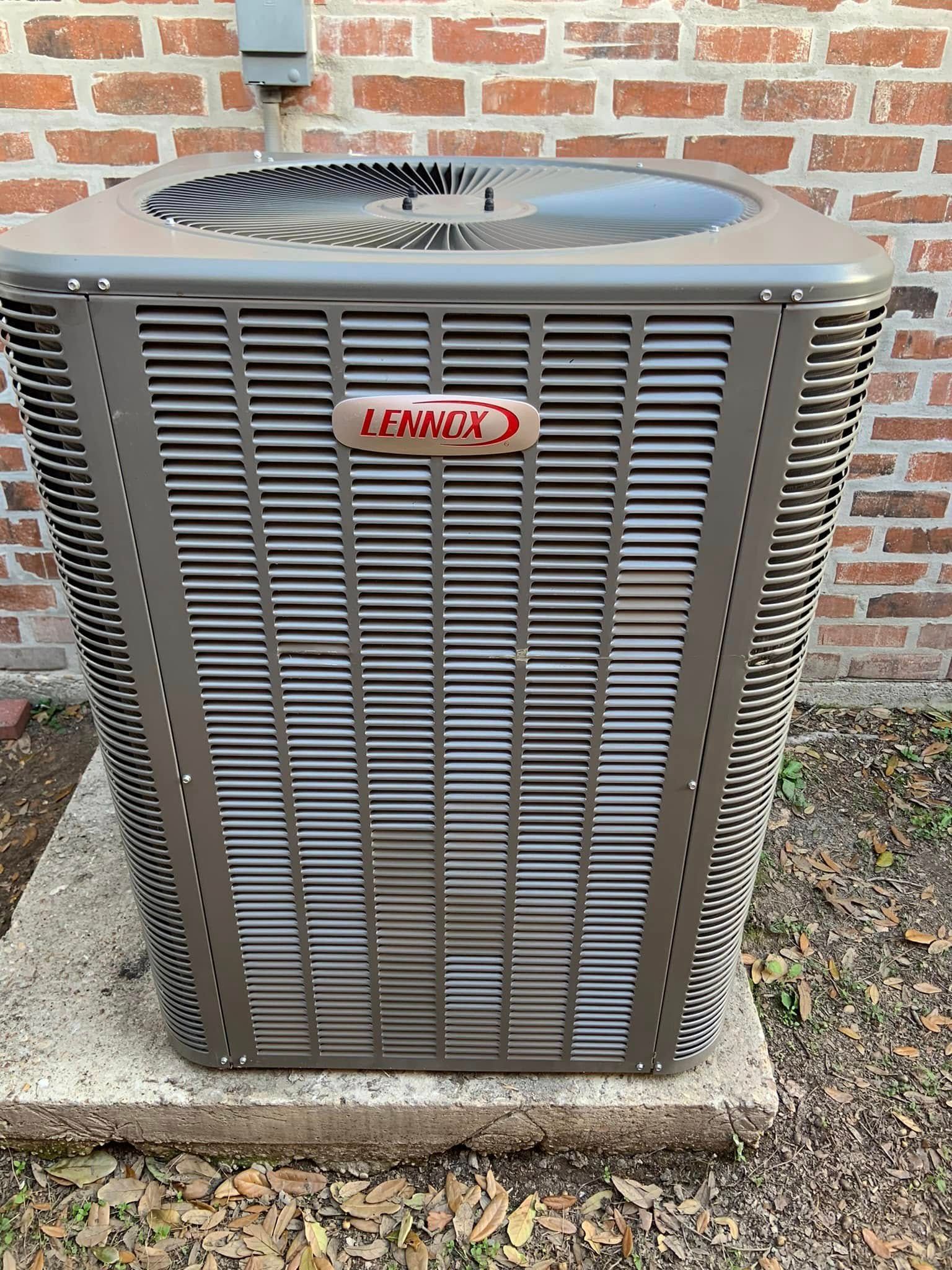 A brown Lennox HVAC condenser unit sits on a concrete pad against a red brick exterior wall.
