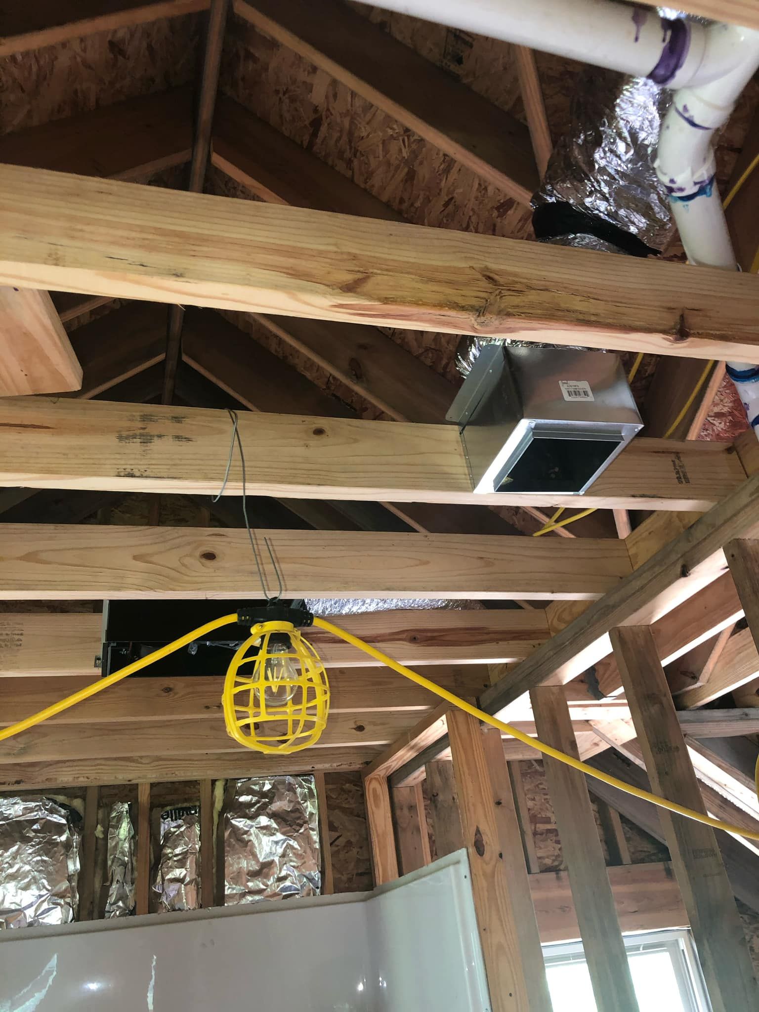 An unfinished wood-framed ceiling with an exposed exhaust fan housing, ductwork, plumbing, and a hanging work light.
