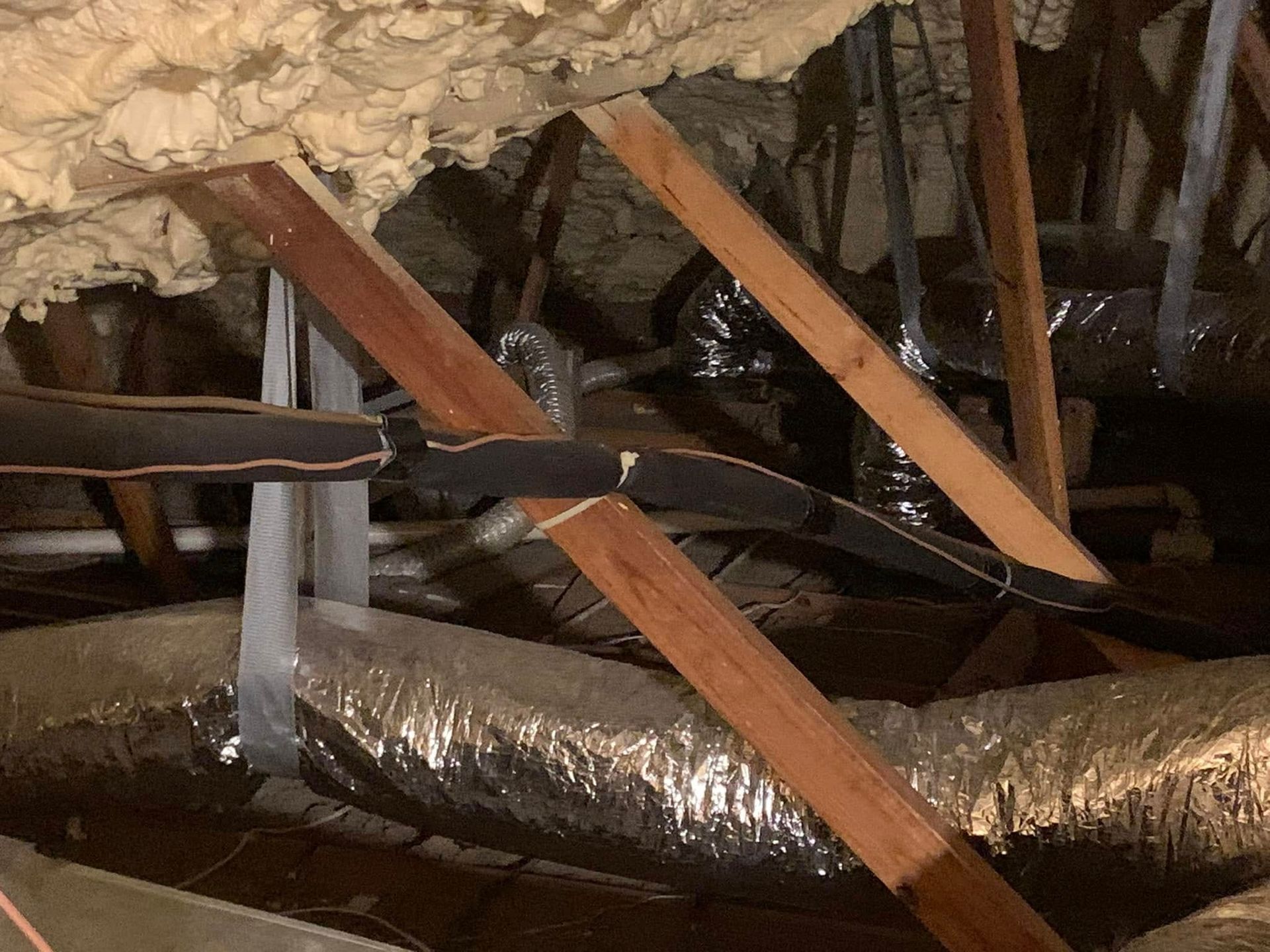 An attic interior showing wooden rafters, spray foam insulation, and flexible silver air ducts supported by gray straps.
