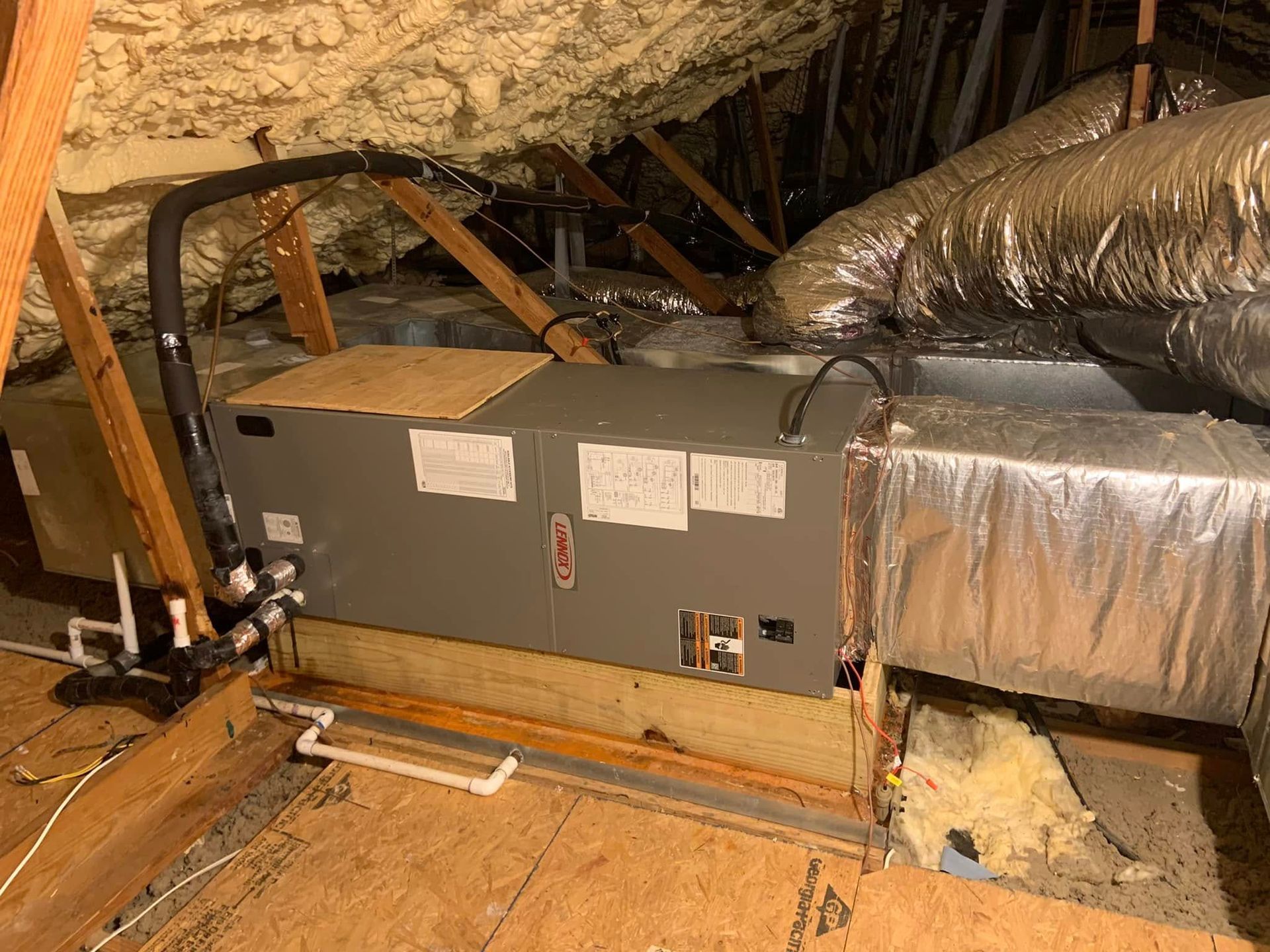 A grey HVAC air handler unit installed in an attic crawl space with foam insulation and foil-wrapped ductwork.