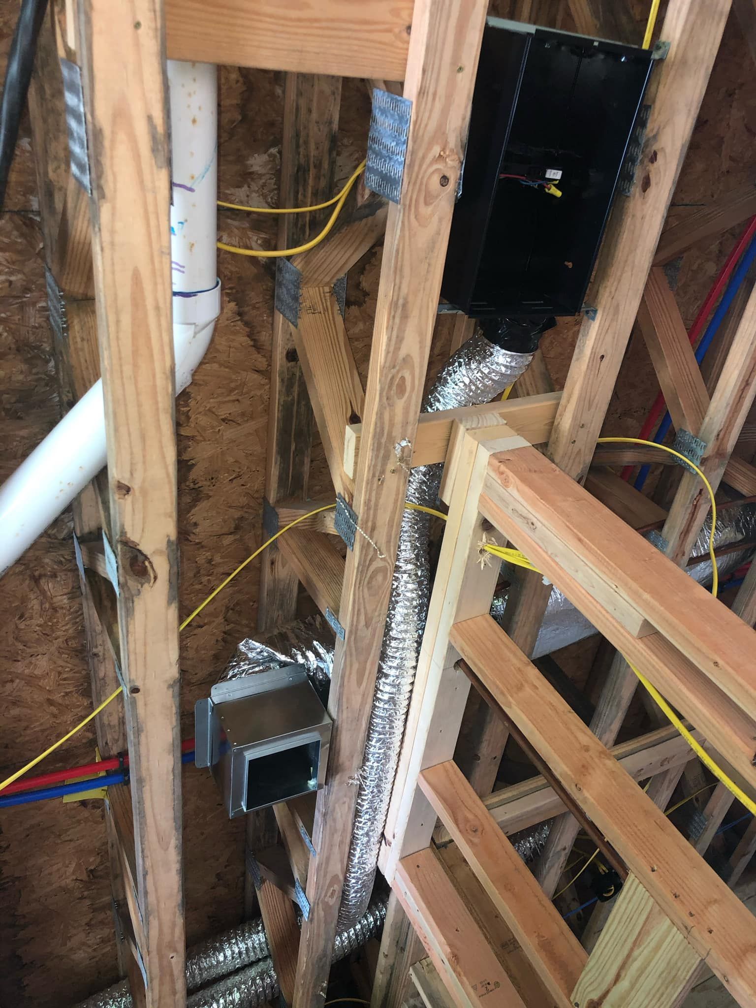 Construction interior showing wooden framing, a black electrical box, silver flexible ductwork, and white PVC piping.