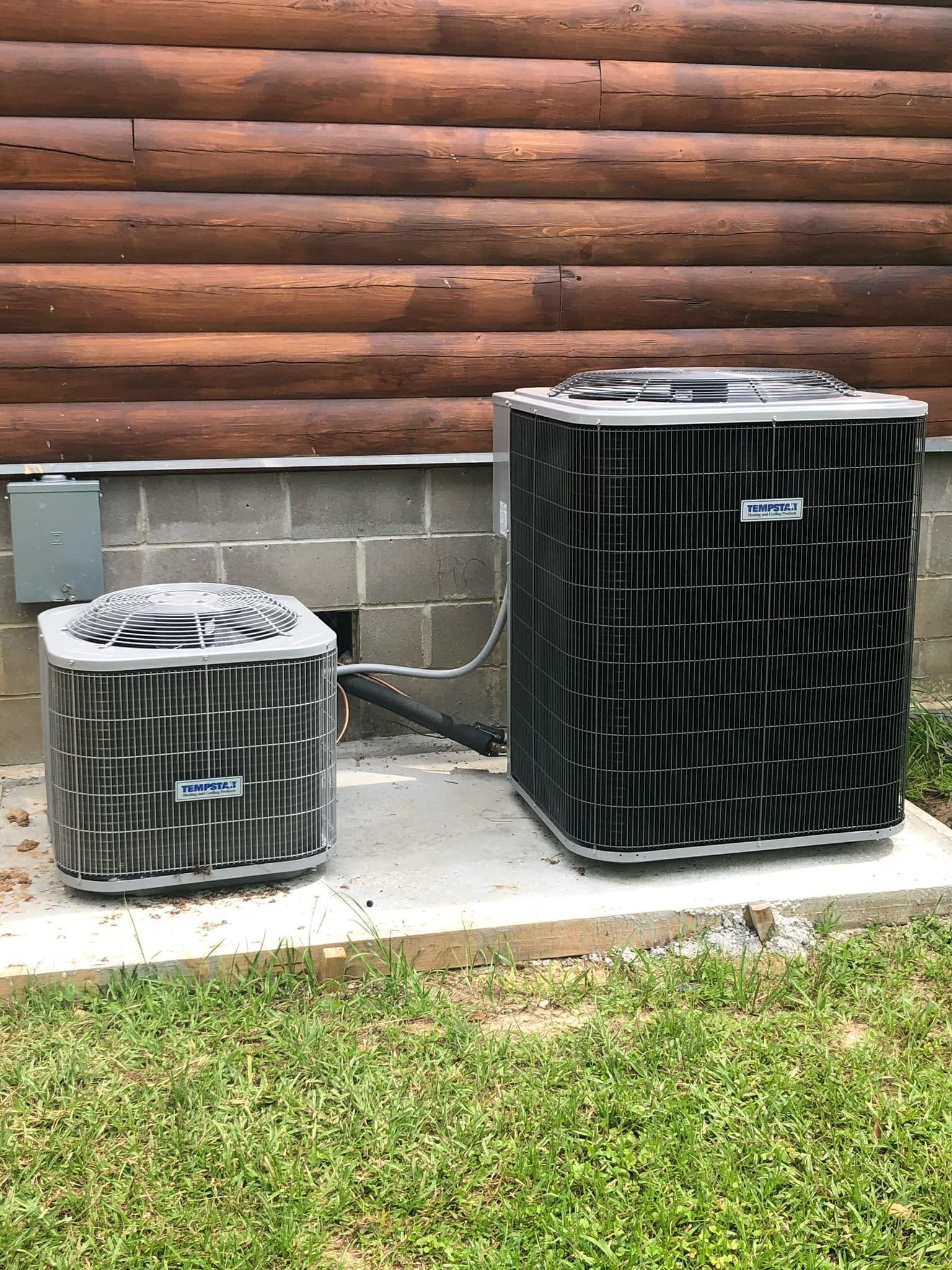 Two air conditioner units sit on a concrete pad against the stone foundation of a log home.