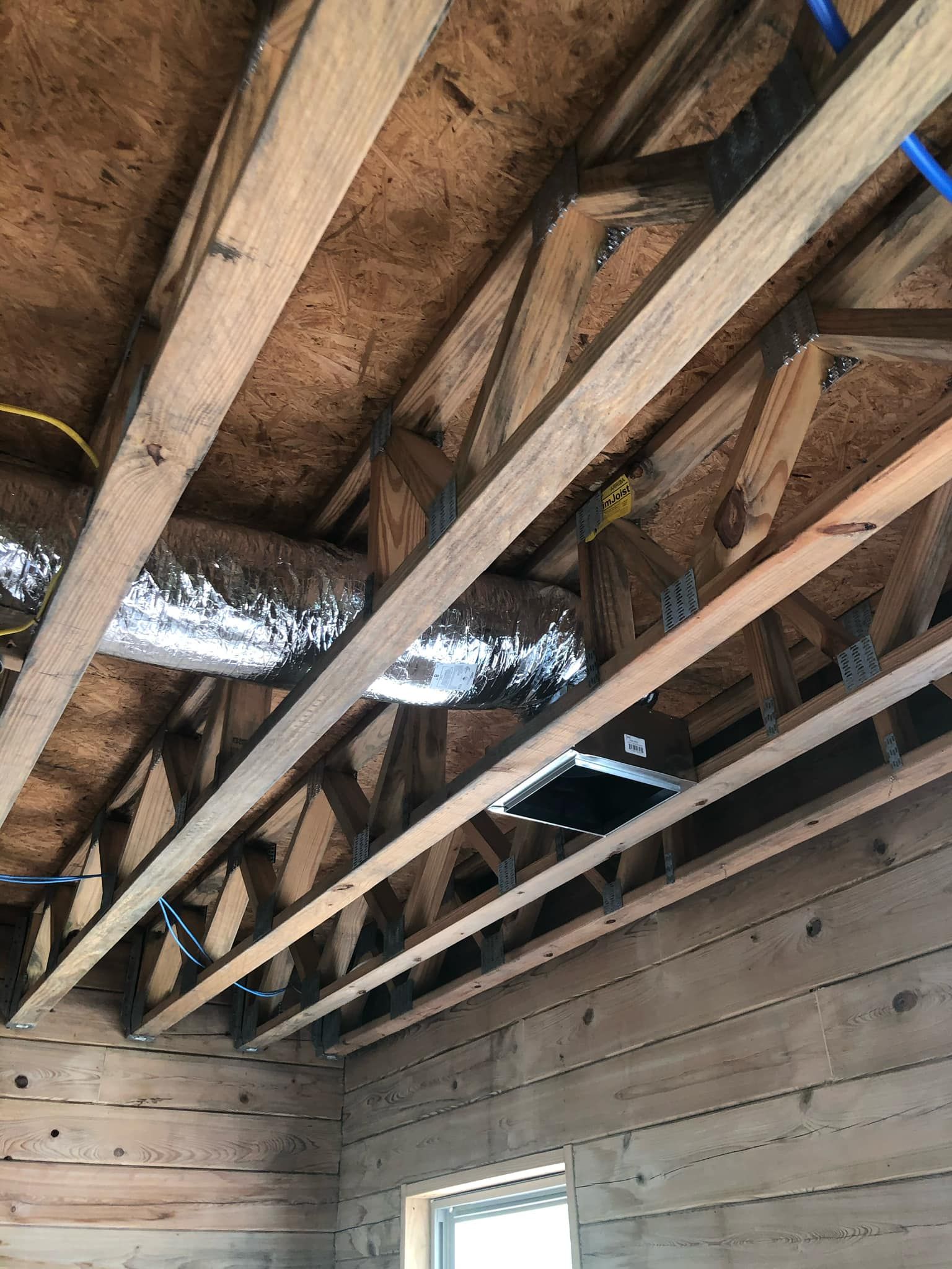 Exposed wooden roof trusses, insulated ductwork, and electrical wiring visible above an unfinished log cabin wall.