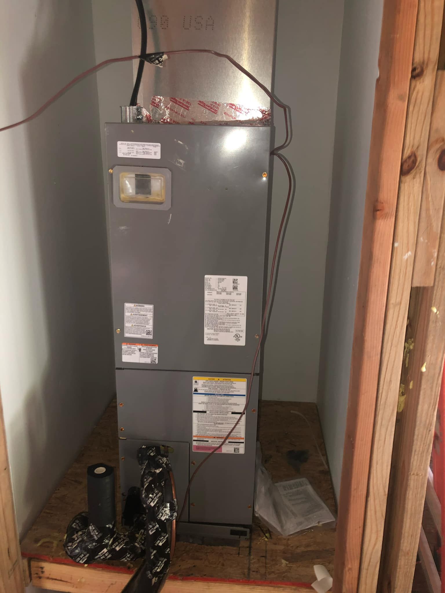 A grey HVAC air handler unit standing in a small, unfinished wooden utility closet with insulated pipes at the base.