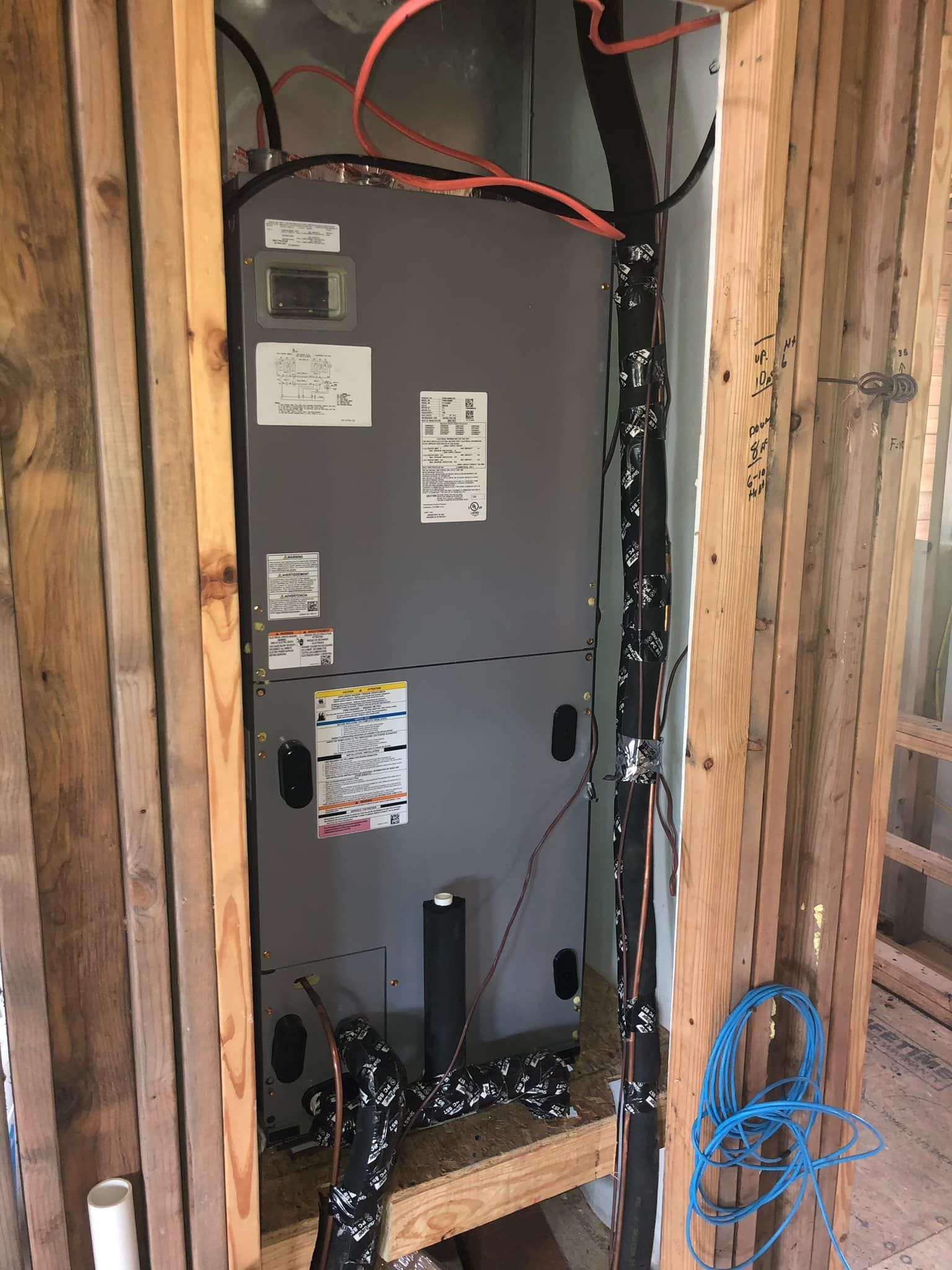 A gray HVAC air handler unit sits inside a framed wooden wall enclosure with exposed wiring and insulated piping.