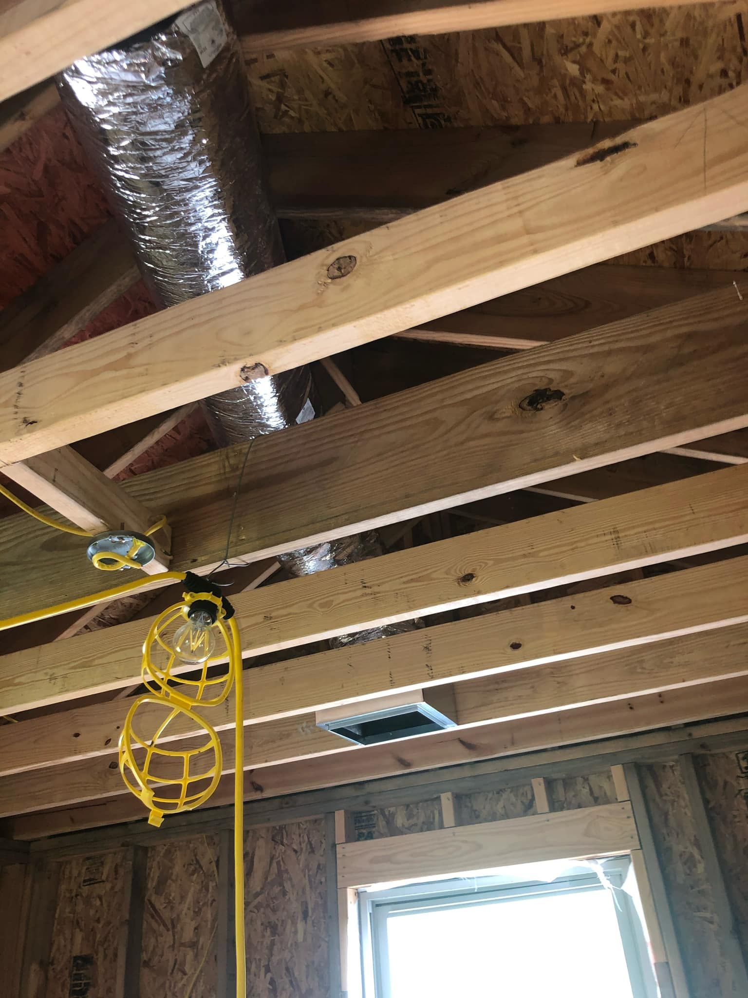 An unfinished room under construction with exposed wooden ceiling joists, metal ductwork, and a hanging yellow work light.
