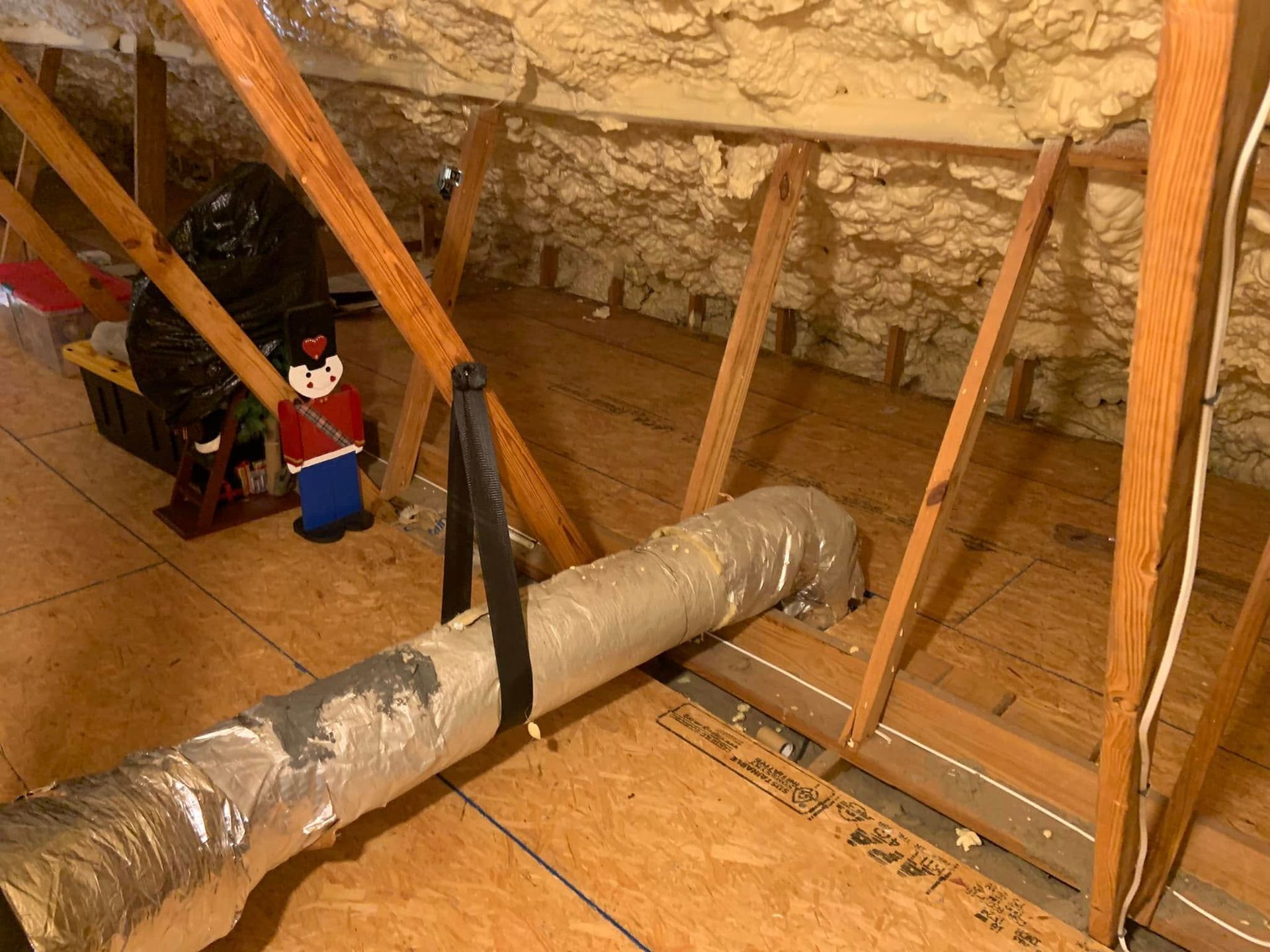 A view inside an attic with wooden rafters, spray foam insulation, flooring, and a suspended silver air duct.