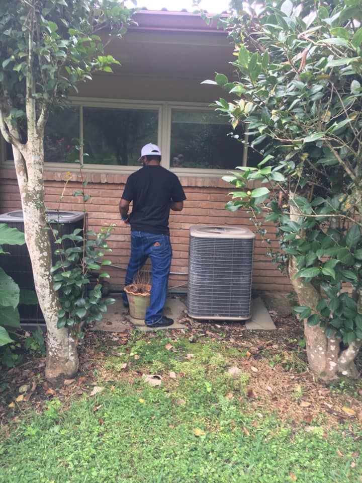 A person stands in a yard by a brick house wall, working on an outdoor air conditioning unit next to a bucket.