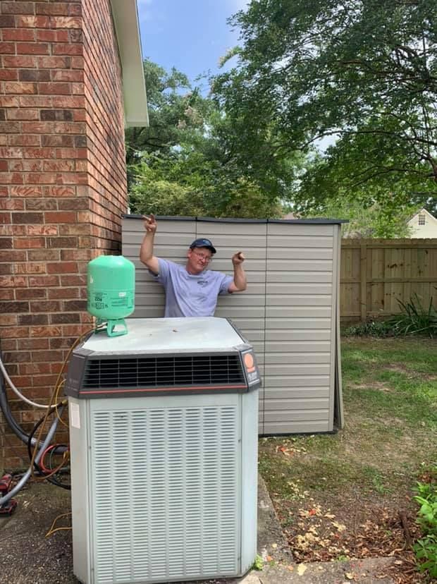 A person in a light shirt and cap stands by a backyard HVAC unit with a refrigerant cylinder on top, smiling at camera.