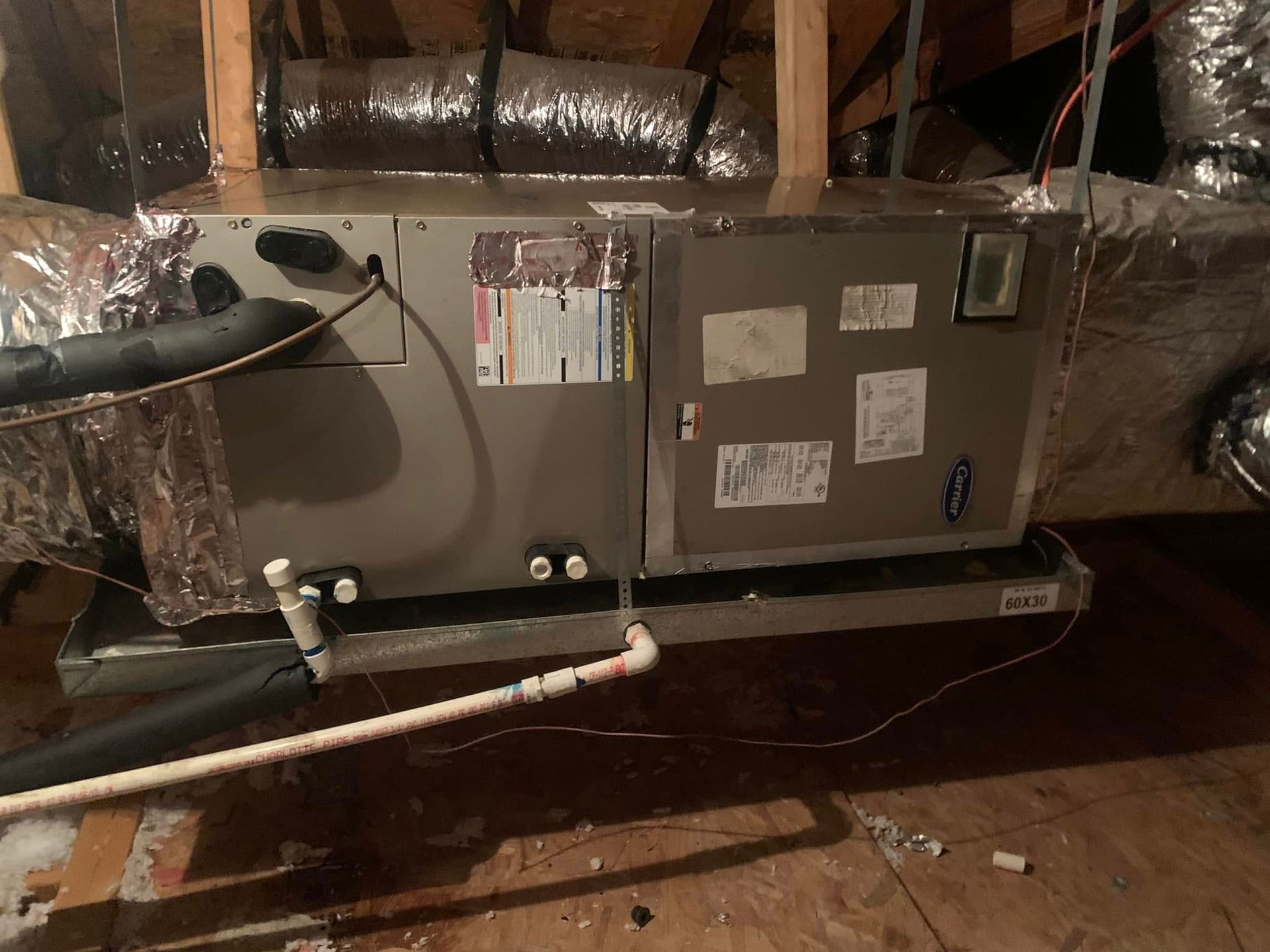 An HVAC air handler unit sits in an attic, featuring metal ducting, a condensate drain pipe, and a safety overflow tray.