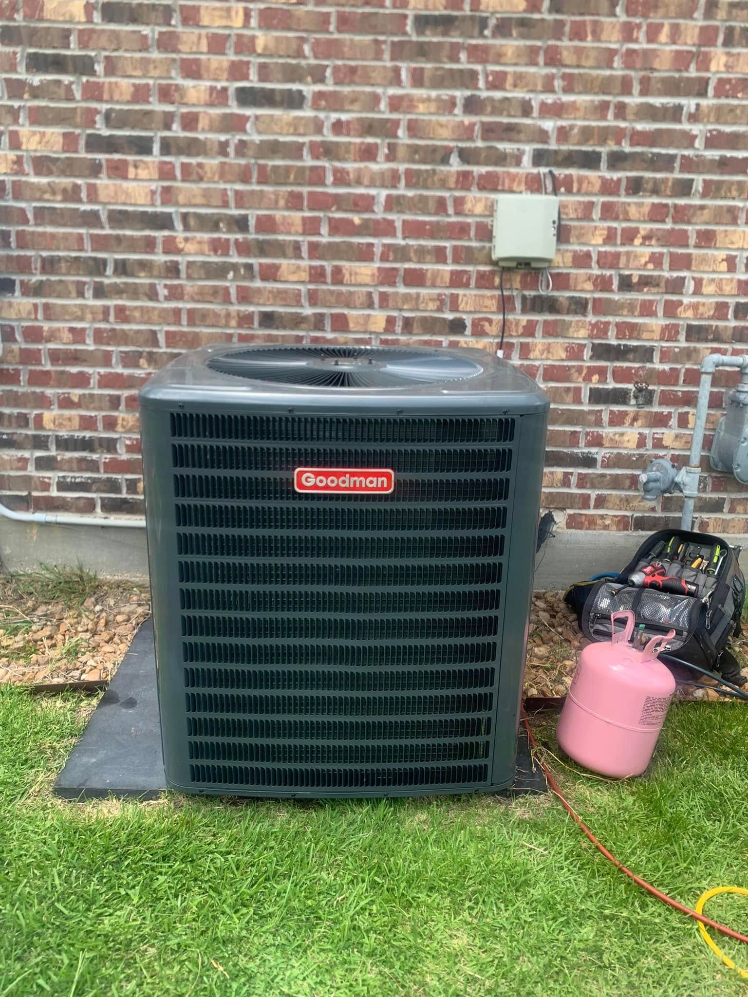 A gray Goodman brand air conditioning unit sits on a pad in front of a brick wall, with a refrigerant tank nearby.