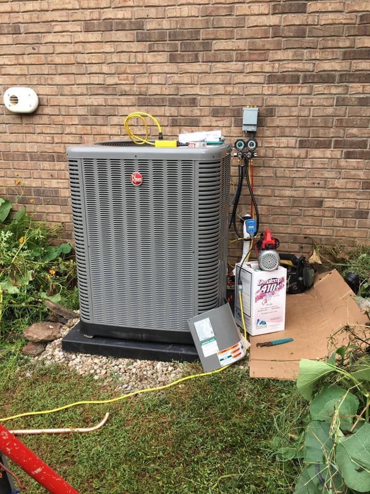 A gray Rheem HVAC condenser unit sits on gravel against a brick wall, undergoing repair with tools and gauges attached.