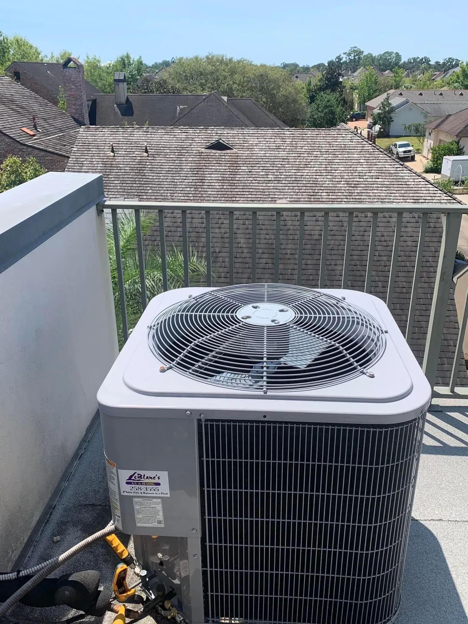 A gray air conditioning unit sits on a rooftop patio with a metal railing, overlooking a neighborhood with shingled roofs.