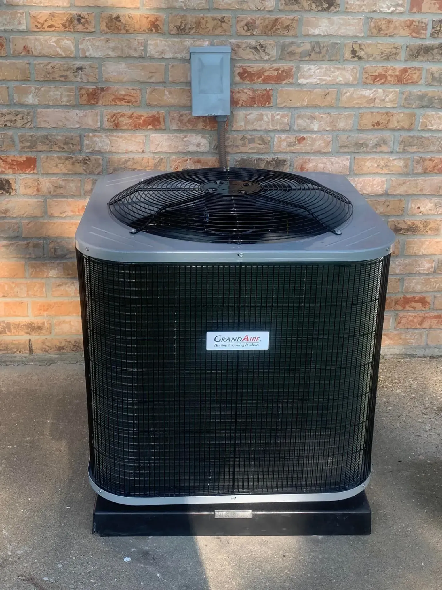 A square outdoor air conditioning unit with a black metal grille, sitting on a concrete pad against a brick wall.