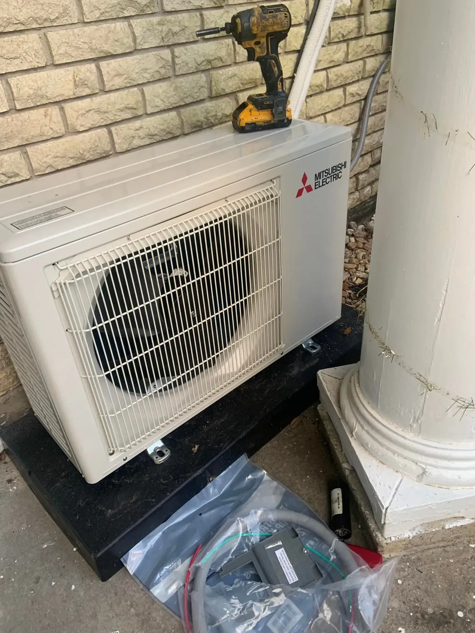 A Mitsubishi heat pump outdoor unit on a black base next to a white column, with a power drill sitting on top.