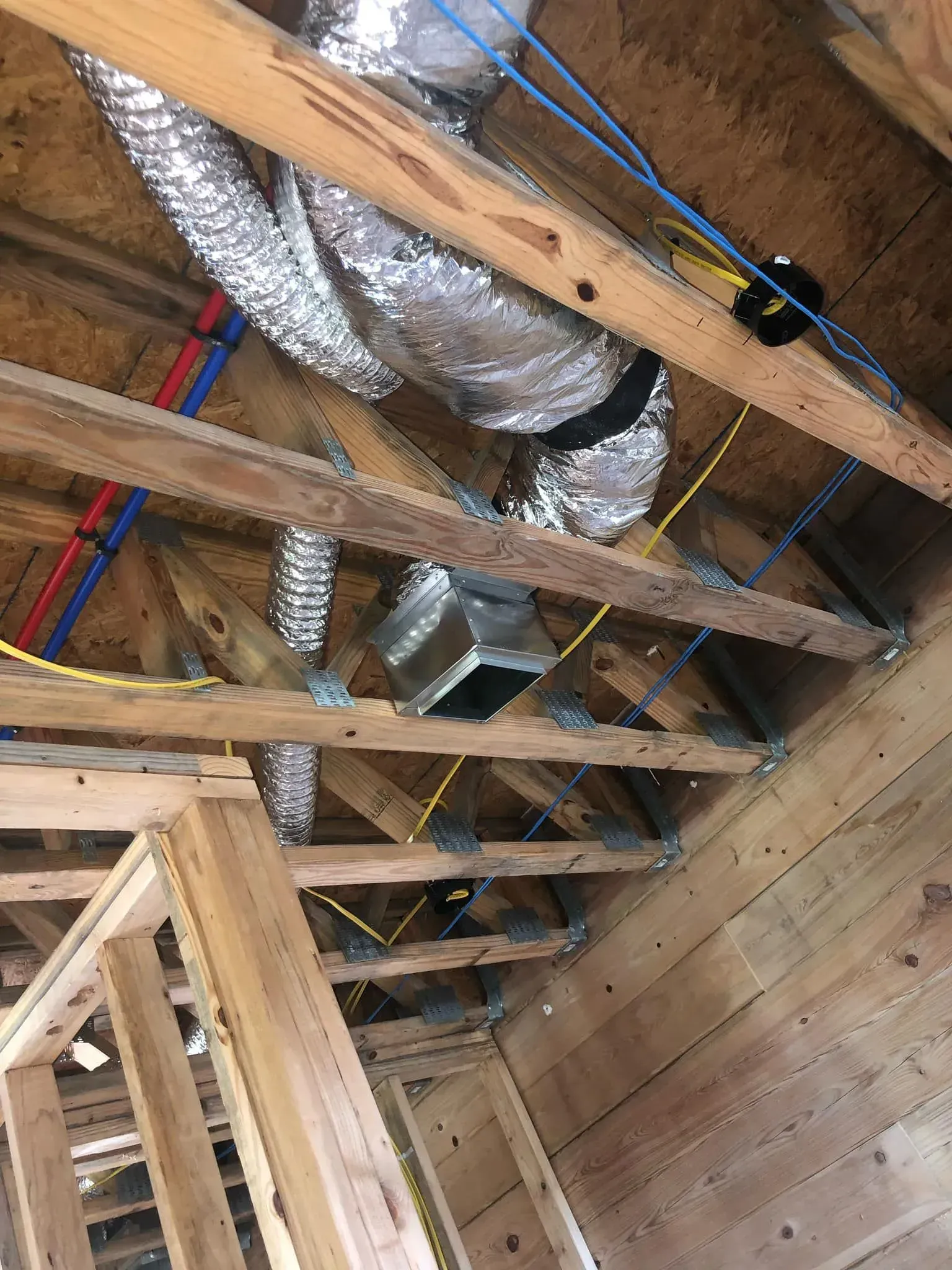 A construction site view of ceiling joists with silver flexible HVAC ducts, red and blue PEX pipes, and yellow wiring.