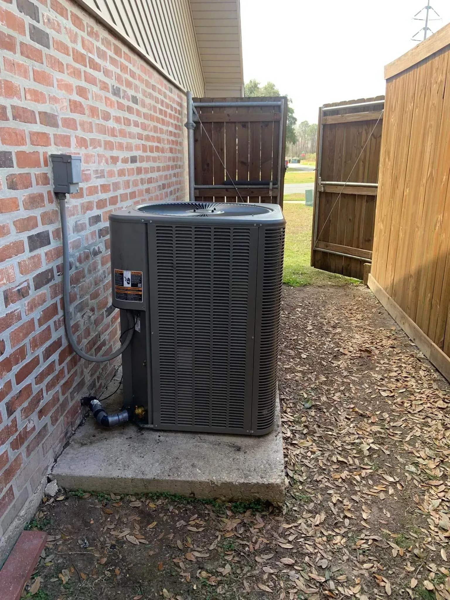 A grey HVAC unit sits on a concrete pad next to a brick wall, situated between two wooden privacy fences.