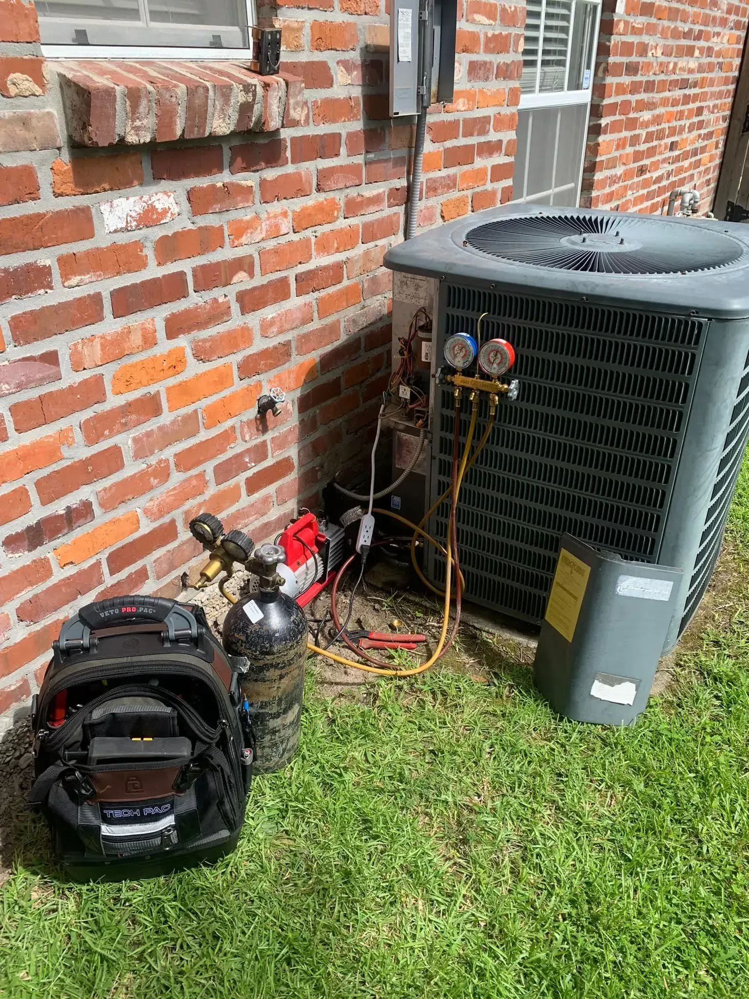 HVAC technician’s gear, including gauges and a tool bag, sits on the grass beside an outdoor air conditioning unit.