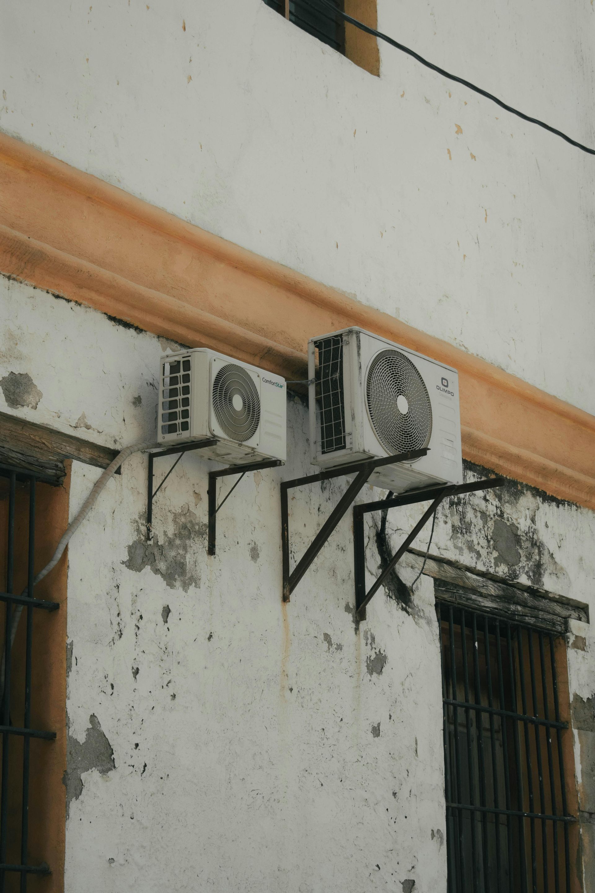 Two white air conditioner units mounted on exterior wall brackets under a horizontal orange ledge on a white building.