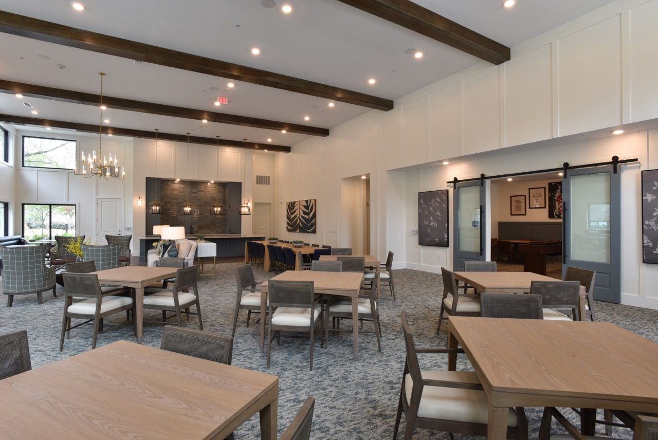 Bright communal lounge with multiple tables, chairs, and a modern kitchen area.