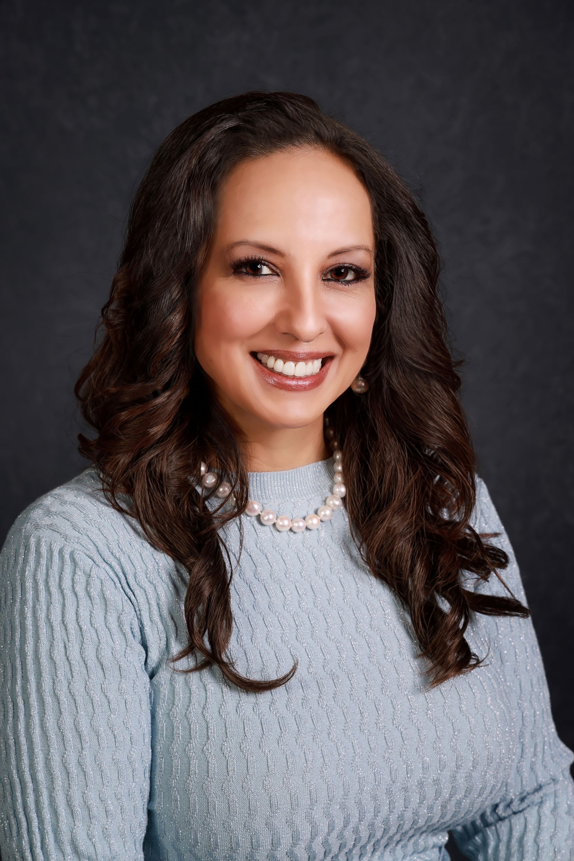 A woman wearing a blue sweater and a pearl necklace is smiling for the camera