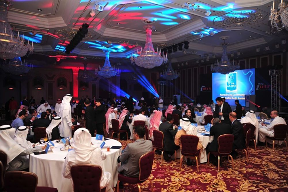 Formal event in a ballroom. Guests seated at round tables. Red and blue stage lighting.