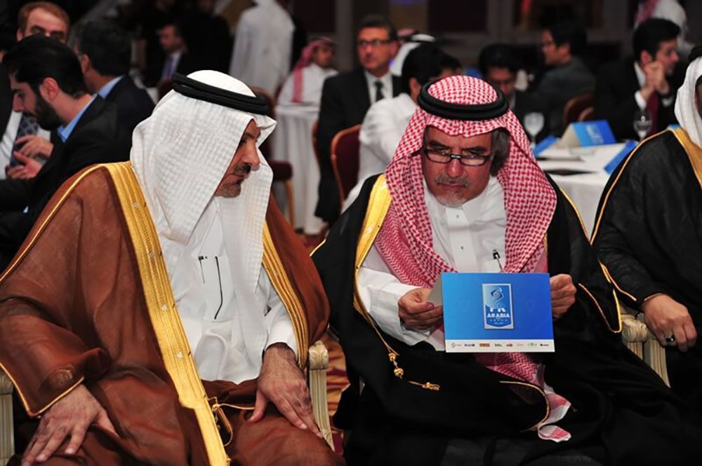 Two men in traditional Arab dress; one holds a tablet. Meeting setting with others.